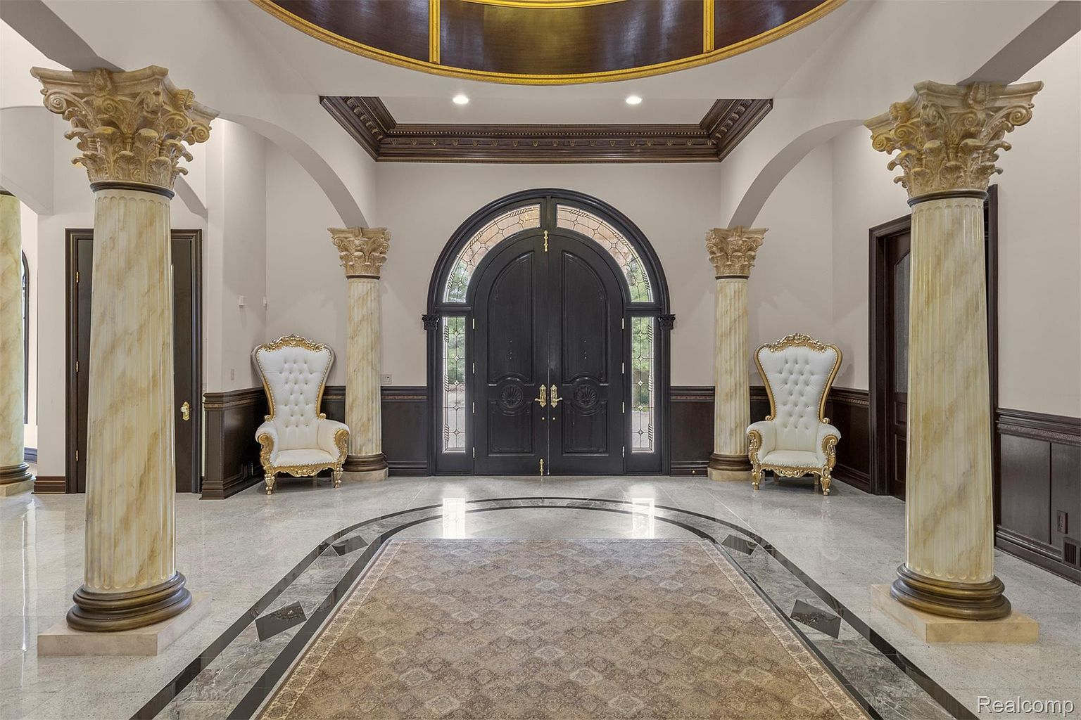 This grand entryway features marble flooring with a decorative rug, flanked by ornate columns and elegant armchairs. A large, arched double door serves as the focal point, complemented by rich wood trim and detailed ceiling work. The space exudes luxury and sophistication, creating a memorable first impression.