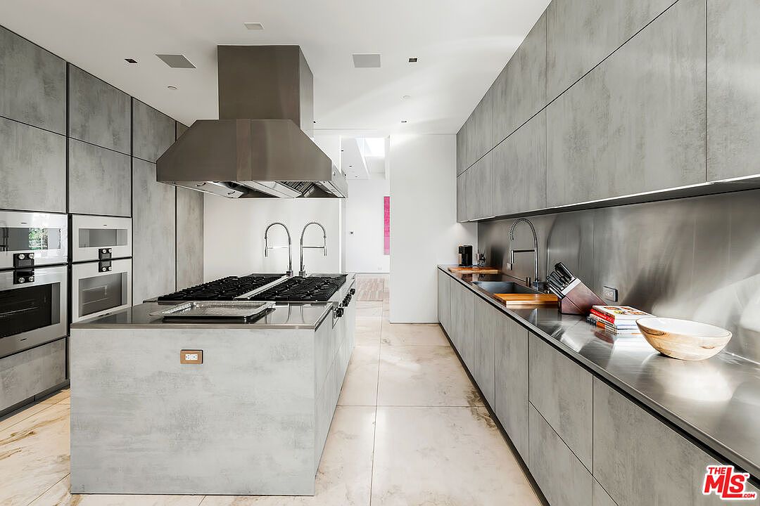 This is a modern kitchen featuring sleek, gray cabinetry and stainless steel countertops. A large island with a cooktop and a stainless steel range hood dominates the center of the room. The overall design is minimalist and industrial, with clean lines and a focus on functionality.
