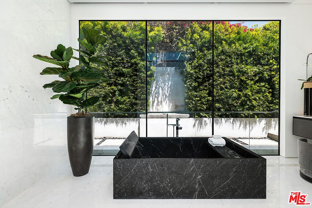 This is a luxurious primary bathroom featuring a modern, dark marble bathtub positioned in front of a large window offering a view of lush greenery. A tall potted plant adds a touch of nature to the interior, while the white marble flooring and walls create a clean and sophisticated ambiance. The overall impression is one of tranquility and high-end design.