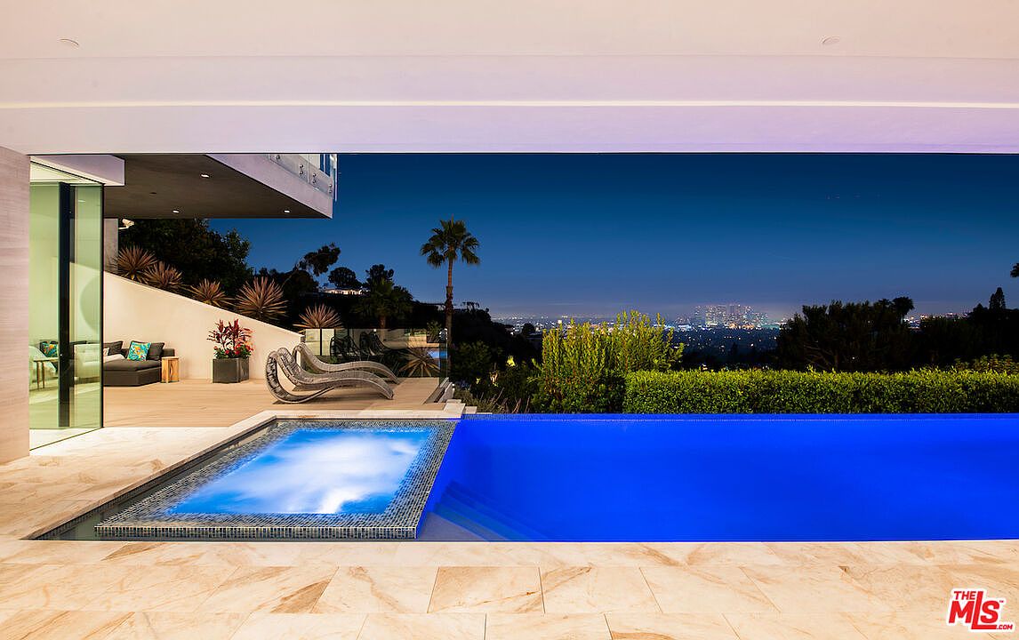 This exterior shot showcases a luxurious pool and spa area at dusk. The pool features vibrant blue water, while the spa is illuminated with a soft glow. The surrounding patio is made of light-colored stone, and the view extends to a cityscape in the distance, creating a serene and upscale ambiance.