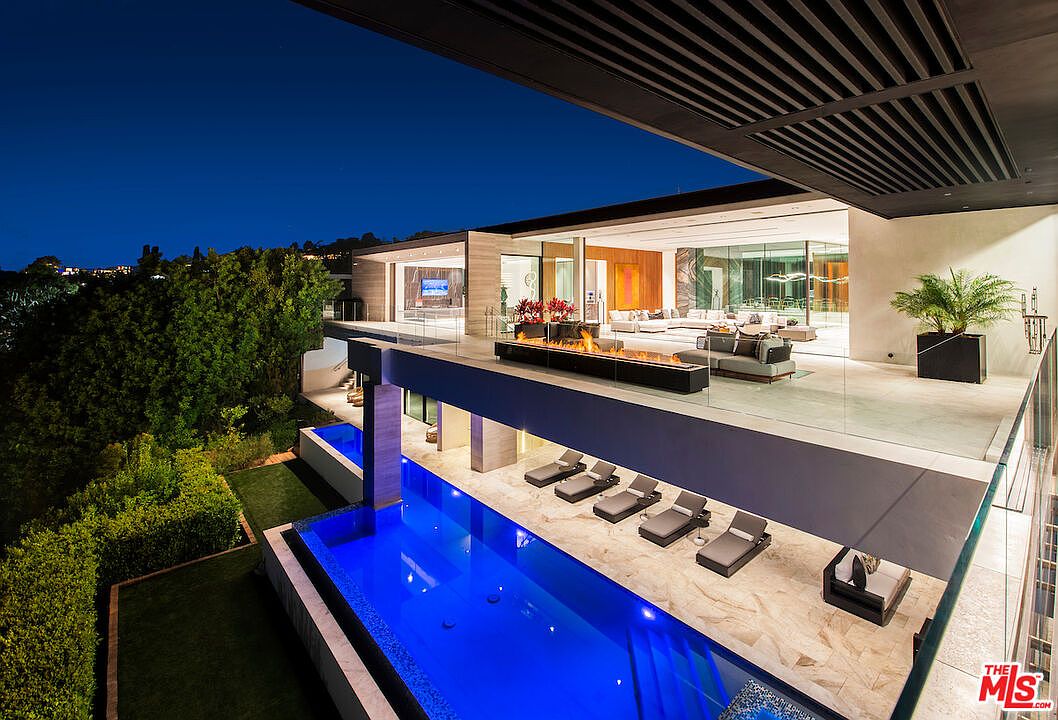 This image showcases a luxurious outdoor space featuring a modern infinity pool with vibrant blue water, surrounded by sleek lounge chairs and a stylish seating area. The pool is situated below an elevated patio with a fire feature and comfortable outdoor furniture, creating a multi-level entertainment area. The architecture is contemporary, with clean lines and glass railings offering unobstructed views of the surrounding landscape.