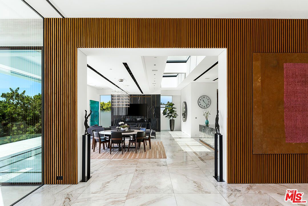 This interior shot showcases a modern living space with an open floor plan, featuring a dining area and a living room. The design incorporates wood paneling, marble flooring, and a neutral color palette, creating a luxurious and airy atmosphere. Large windows and skylights provide ample natural light, enhancing the sense of spaciousness.