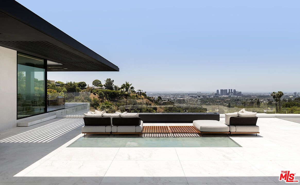 This image showcases a luxurious patio/deck area with modern outdoor furniture, including a modular sofa and a wooden platform. The space features sleek white tiling and a glass-enclosed section, offering stunning panoramic views of the city skyline and surrounding hills. The architectural design is contemporary, emphasizing clean lines and open space, perfect for outdoor relaxation and entertainment.