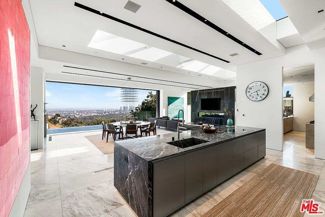 This is a bright, modern kitchen with a large island featuring a dark marble countertop and sleek gray cabinetry. The space is open to the dining and living areas, with a stunning city view visible through a large window. The kitchen also features a black marble backsplash and a wall-mounted television, adding a touch of luxury.