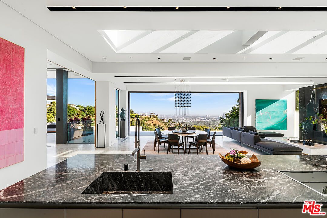 This interior shot showcases a modern, open-concept kitchen and dining area with a luxurious feel. The kitchen features a dark marble countertop with a sink and sleek cabinetry, while the dining area boasts a round table surrounded by chairs, set against a backdrop of a panoramic city view through large windows. The space is bright and airy, enhanced by natural light and a contemporary chandelier.