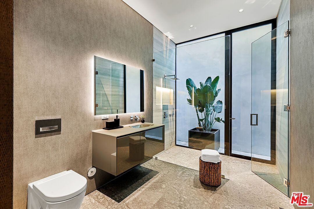 This is a modern primary bathroom featuring a floating vanity with a vessel sink and a large, illuminated mirror. The bathroom has a walk-in shower with glass doors and a view of an outdoor plant area, creating a spa-like atmosphere. The color palette is neutral, with textured walls and stone flooring, contributing to a luxurious and serene ambiance.