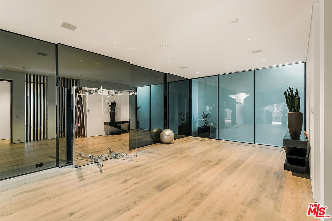This is a spacious home gym featuring light wood flooring, mirrored walls, and large windows that provide ample natural light. The room includes exercise balls and a weight training machine, creating a dedicated space for fitness. The overall impression is modern and well-lit, perfect for a health-conscious buyer.