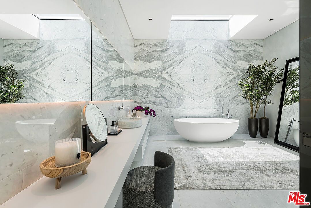 This is a luxurious primary bathroom featuring marble walls and flooring, creating a spa-like atmosphere. A freestanding oval bathtub sits on a light gray rug, complemented by a modern vanity with a vessel sink and a round mirror. The room is illuminated by natural light from skylights, enhancing the clean and elegant design.