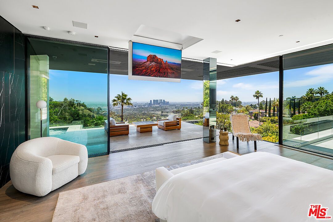 This is a luxurious primary bedroom featuring floor-to-ceiling glass walls that offer panoramic views of the city and surrounding landscape. The room includes a large bed, a comfortable armchair, and a unique ceiling-mounted television. The open layout seamlessly connects the interior with the outdoor scenery, creating a serene and modern living space.