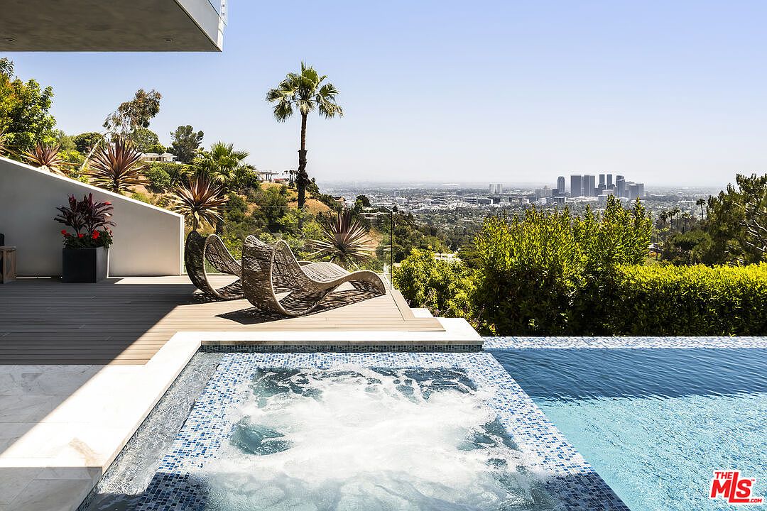 This image showcases a luxurious outdoor pool and spa area with a stunning city view. The foreground features a tiled spa with bubbling water, adjacent to a clear blue swimming pool. Two modern lounge chairs sit on a wooden deck overlooking the cityscape, creating an inviting and relaxing atmosphere.