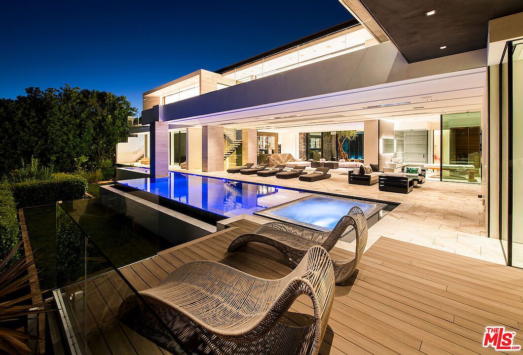 This image showcases a luxurious outdoor pool and spa area of a modern home at dusk. The pool's blue water contrasts with the warm tones of the wooden deck and lounge chairs. The open-concept design seamlessly blends indoor and outdoor living spaces, highlighting the property's entertainment potential.