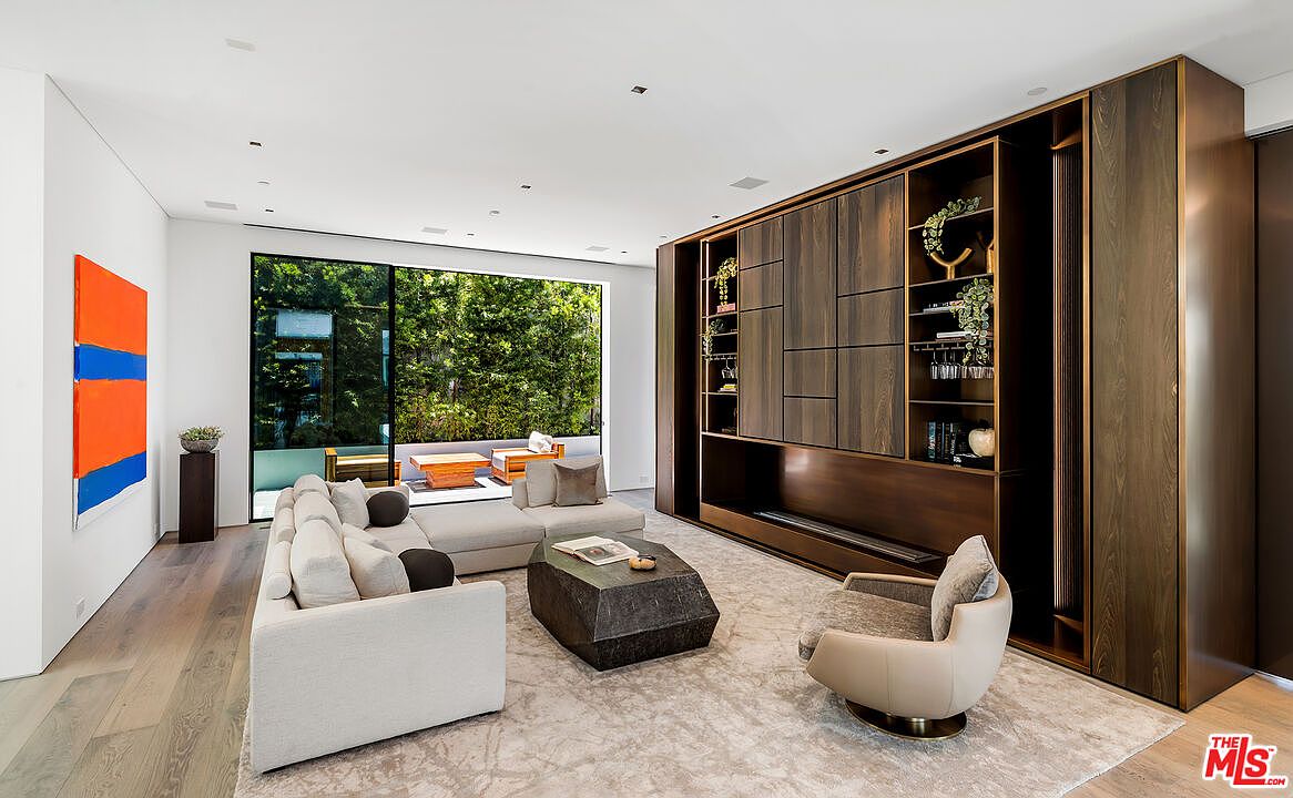 This is a modern living room featuring a large sectional sofa, a unique geometric coffee table, and a custom-built media center with wood paneling. A large window provides ample natural light and a view of the greenery outside. The room is decorated with contemporary art and has a neutral color palette, creating a sophisticated and inviting atmosphere.
