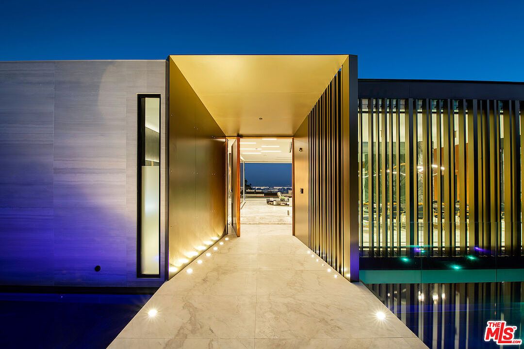 The image showcases a modern home's entryway at dusk, featuring a long, tiled walkway leading to an open doorway. The entrance is framed by gold-toned walls and vertical slats, with subtle lighting illuminating the path. A glimpse of the interior and the outdoor view beyond adds depth and allure, suggesting a luxurious and inviting atmosphere.