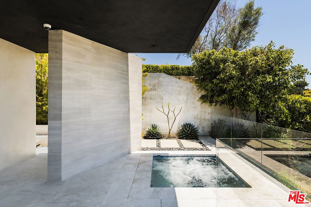 This exterior shot showcases a modern spa area with clean lines and minimalist design. The spa is surrounded by light-colored stone tiles and features a glass railing, offering views of the surrounding greenery. The architectural elements, including the dark overhang and light-colored walls, create a sophisticated and serene atmosphere.