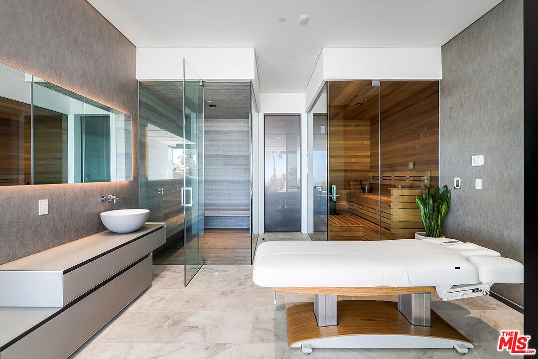 This is a luxurious primary bathroom featuring a modern design with a spa-like atmosphere. The room includes a sleek vanity with a vessel sink, a large mirror with integrated lighting, a glass-enclosed shower, and a sauna. A massage table is positioned in the foreground, suggesting a focus on relaxation and wellness.