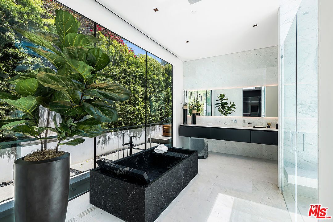 This is a luxurious primary bathroom featuring a modern black marble bathtub, a sleek floating vanity with a large mirror, and a glass-enclosed shower. The room is bright and airy, with a large window offering a view of lush greenery, and the marble flooring adds to the sophisticated aesthetic. A large potted plant adds a touch of nature to the space.