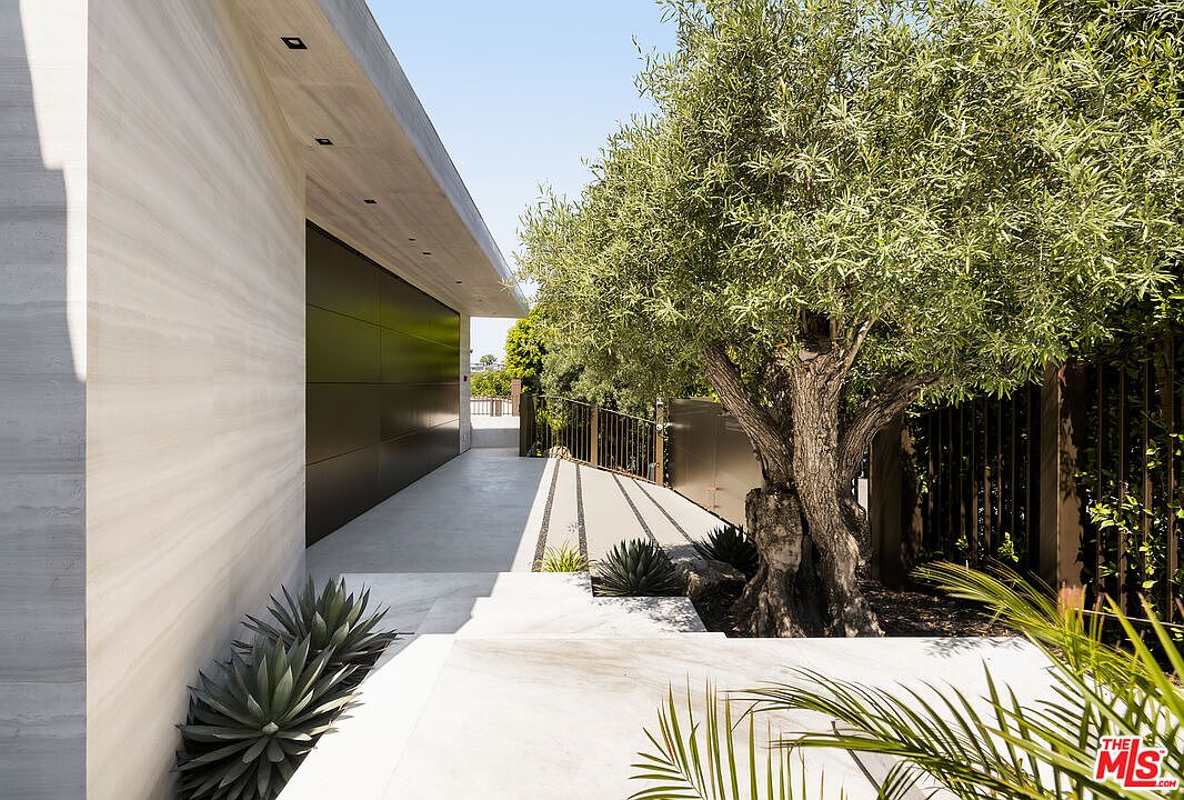 This image showcases a modern home's entryway, featuring a sleek, minimalist design with clean lines and a neutral color palette. The exterior is characterized by a light-colored stone wall, a dark-paneled garage door, and a concrete walkway leading to the entrance. A mature olive tree and other carefully placed greenery add a touch of nature and sophistication to the property's curb appeal, creating an inviting and stylish first impression.