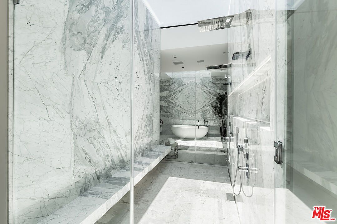 This is a luxurious primary bathroom featuring extensive marble walls and flooring, creating a spa-like atmosphere. A freestanding bathtub is visible in the background, and a glass-enclosed shower area with modern fixtures is in the foreground. The design emphasizes clean lines and a bright, open space, suggesting high-end finishes and a focus on relaxation.