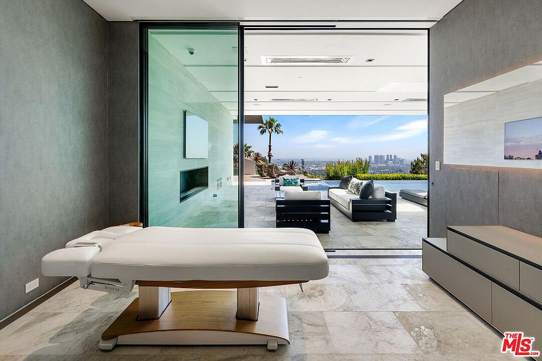 This interior shot showcases a modern home gym with a massage table as a focal point. Large sliding glass doors open to an outdoor patio with city views, creating a seamless indoor-outdoor transition. The room features sleek gray walls, marble flooring, and contemporary built-in storage, contributing to a luxurious and serene atmosphere.