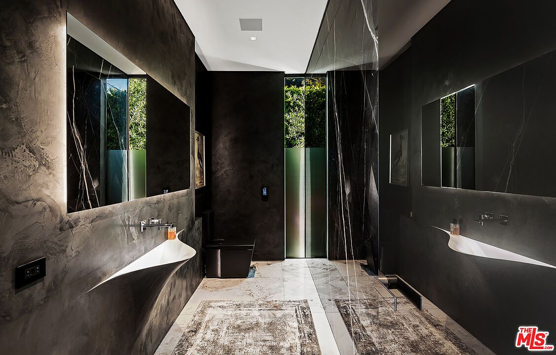 This is a modern primary bathroom featuring dark, textured walls and marble accents. The room includes a unique, sculptural sink design, a large mirror with integrated lighting, and a glass-enclosed shower area. The overall aesthetic is luxurious and contemporary, with a focus on high-end materials and design.