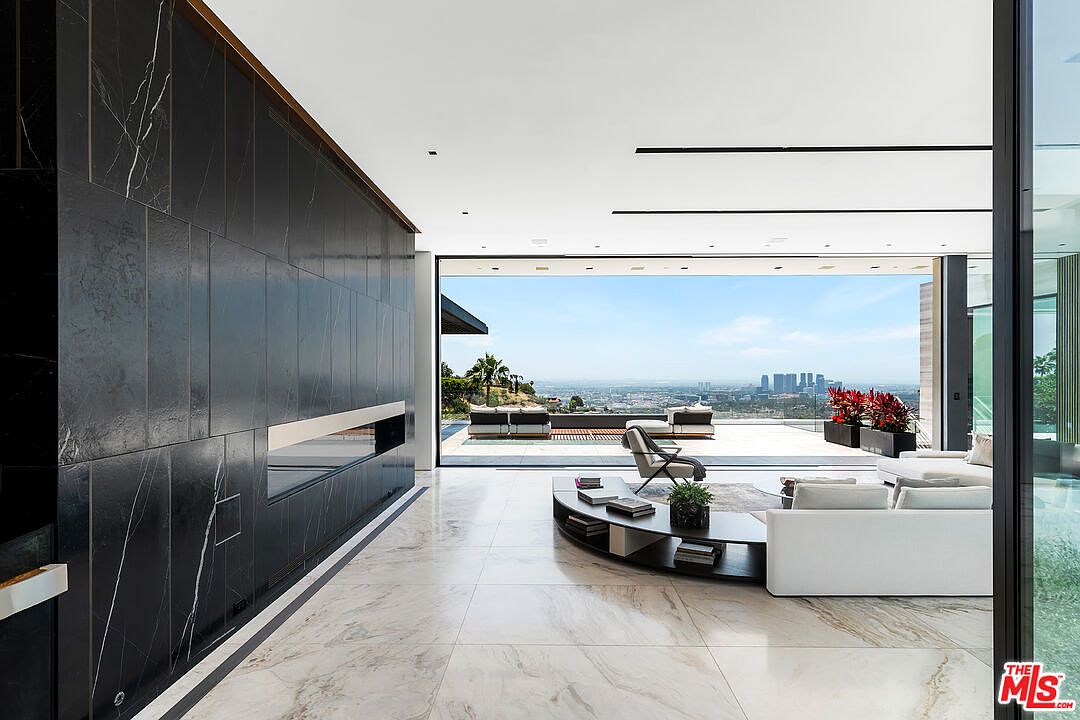 This is a luxurious living room featuring a sleek, modern design with floor-to-ceiling windows offering panoramic city views. The room includes a white sofa, a unique curved coffee table, and a stylish accent chair, all set on a light-colored marble floor. A striking black marble wall with a built-in fireplace adds a dramatic focal point to the space.