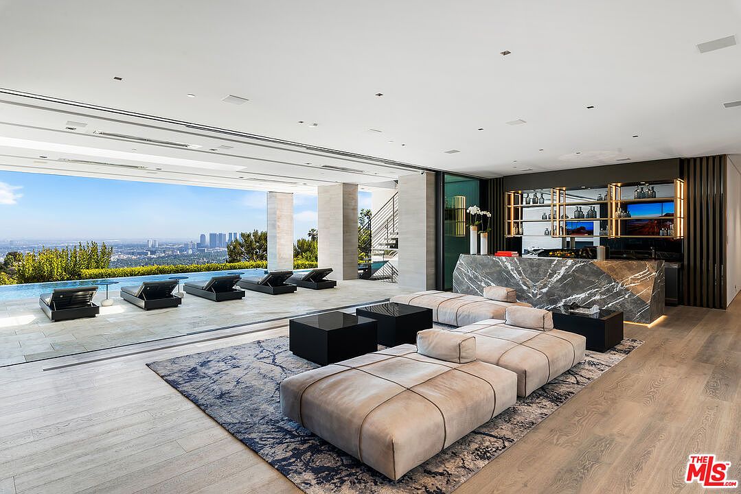 This is a luxurious living room with an open floor plan that seamlessly blends indoor and outdoor spaces. The room features modern furniture, including a large sectional sofa and sleek black coffee tables, set upon a stylish area rug. A bar area with marble countertops and illuminated shelving adds a touch of sophistication, while floor-to-ceiling windows offer stunning views of the city skyline and a nearby pool.