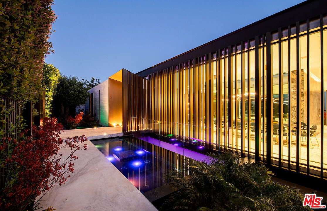 This image showcases a modern home's exterior with a sleek pool and spa area. The architecture features clean lines, vertical wooden slats, and large windows that offer a glimpse into the interior. The pool is illuminated with blue lights, creating a luxurious and inviting atmosphere, complemented by lush landscaping.