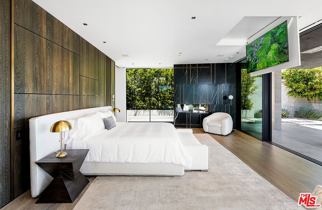 This is a luxurious primary bedroom featuring a large bed with white linens, a dark wood accent wall, and a modern fireplace. The room has floor-to-ceiling windows offering views of the outdoors, and a retractable television is mounted on the ceiling. The overall style is contemporary and sophisticated, creating a serene and upscale atmosphere.
