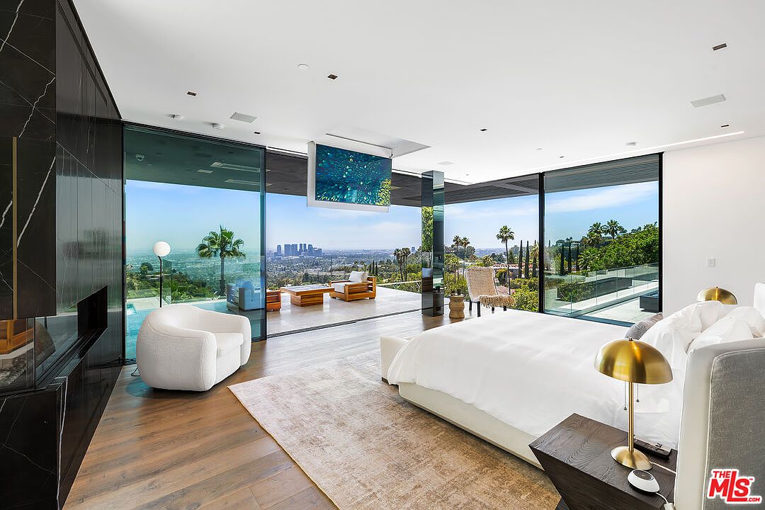 This is a luxurious primary bedroom featuring floor-to-ceiling glass walls that offer panoramic views of the city and surrounding landscape. The room includes a large bed with white linens, a modern fireplace, and a comfortable seating area. The design emphasizes natural light and seamless indoor-outdoor living, creating a serene and upscale atmosphere.