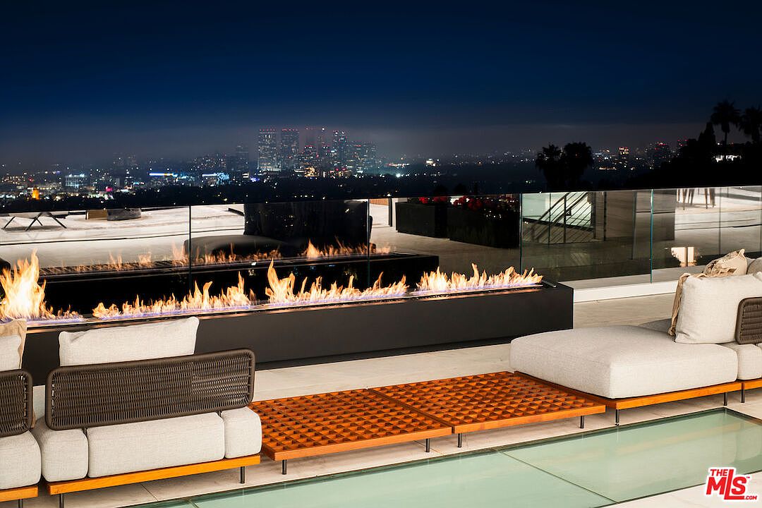This image showcases a luxurious outdoor patio or balcony area at night, featuring a modern fire pit, comfortable lounge seating, and a glass-paneled floor. The cityscape in the background adds to the upscale ambiance, suggesting a high-end property with stunning views. The perspective is cinematic, emphasizing the depth and breadth of the outdoor space.
