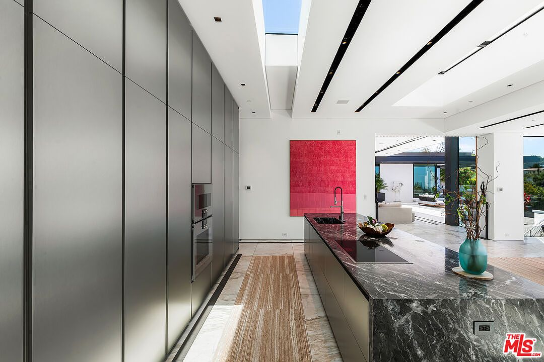 This is a modern kitchen with sleek, gray cabinetry and a dark marble countertop island. A large red painting adds a pop of color to the white walls. The kitchen features a skylight and opens to a living area, creating an open and airy space.