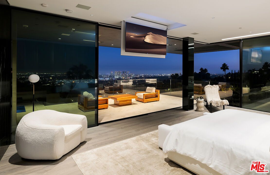 This is a luxurious primary bedroom featuring floor-to-ceiling glass walls that offer panoramic city views. The room includes a large bed with white linens, a cozy armchair, and a modern entertainment system with a ceiling-mounted television. The design emphasizes open space and seamless indoor-outdoor living, creating a sophisticated and relaxing atmosphere.