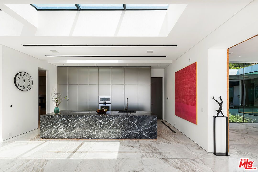 This is a bright and modern kitchen featuring a large marble island with a sink and faucet. The kitchen has sleek, stainless steel cabinetry and appliances, with a large skylight providing ample natural light. A red artwork hangs on the wall, and a sculpture stands on a pedestal, adding an artistic touch to the space.