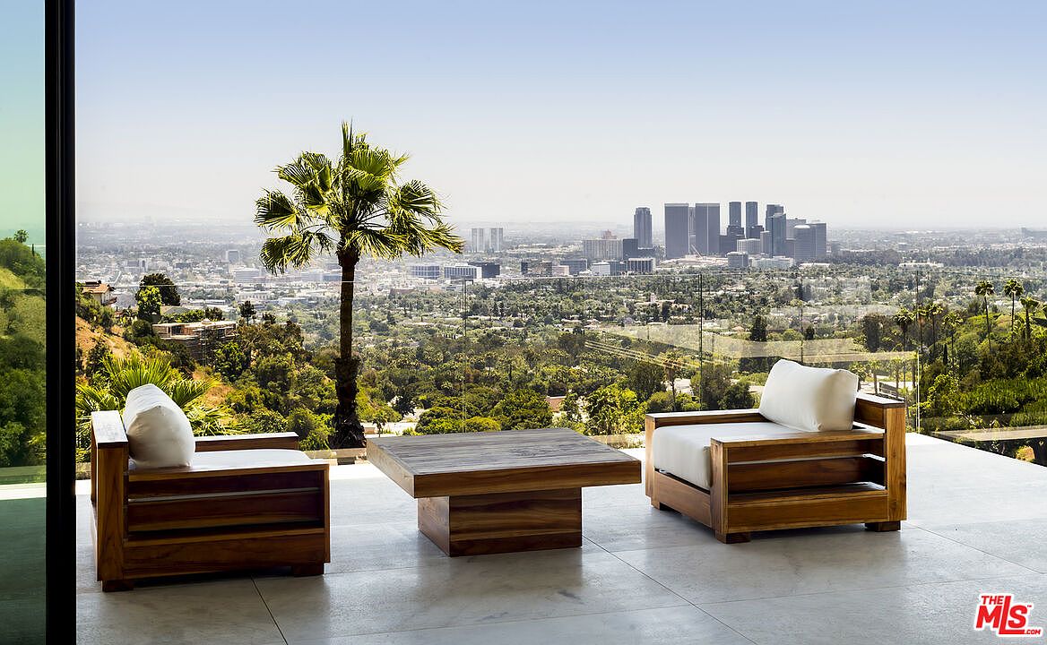 This image showcases a luxurious outdoor patio or balcony area with stunning city views. The space is furnished with modern wooden chairs and a matching coffee table, creating a comfortable and stylish seating arrangement. The backdrop features a sprawling cityscape, lush greenery, and a clear blue sky, enhancing the property's appeal and offering a sense of tranquility and exclusivity.