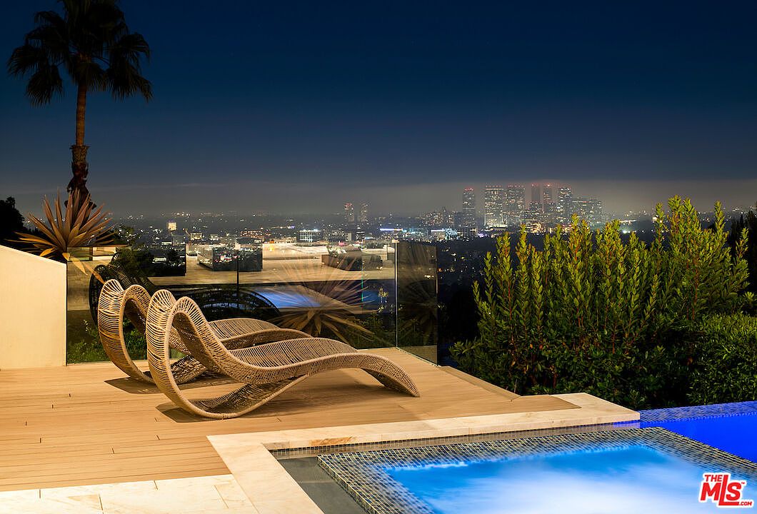 This image showcases a luxurious outdoor pool area at night, featuring two modern lounge chairs on a wooden deck overlooking a cityscape. The pool's edge is tiled with mosaic patterns, and lush greenery surrounds the deck, enhancing the sense of privacy and tranquility. The scene is illuminated by the city lights and the pool's underwater lighting, creating a sophisticated and inviting ambiance.