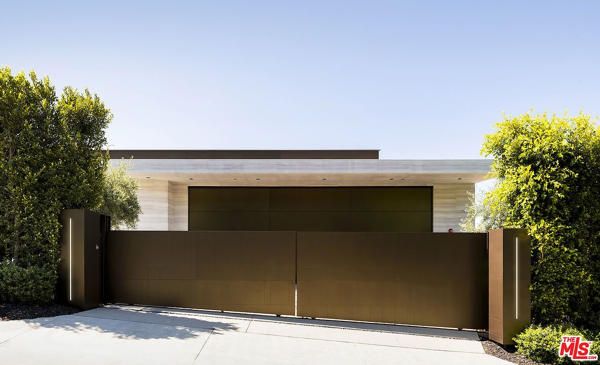 The image showcases a modern home's entryway, featuring a large, dark-colored gate with a geometric pattern. Flanking the gate are two dark pillars with vertical light strips, adding a touch of contemporary design. Lush greenery surrounds the entrance, and the house itself has a flat roof and a light-colored facade, creating a sleek and inviting first impression.