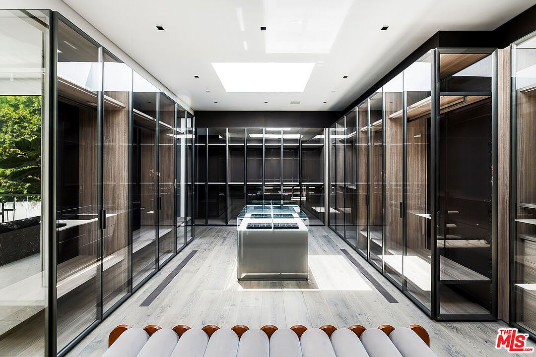 This is a luxurious walk-in closet featuring glass-fronted wardrobes with wooden interiors and black frames. A central island with a glass top provides additional storage and display space. The room is well-lit with a skylight and recessed lighting, creating a bright and sophisticated atmosphere.