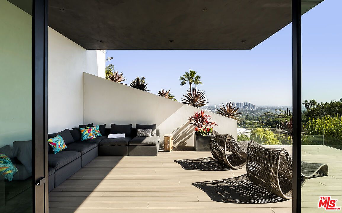 This image showcases a stylish outdoor patio or balcony area, featuring a modern sectional sofa with decorative pillows and two unique woven chairs. The space is designed with a sleek, minimalist aesthetic, complemented by a wooden deck and a low wall adorned with various plants. The view extends to a cityscape in the distance, creating a luxurious and inviting outdoor living space.