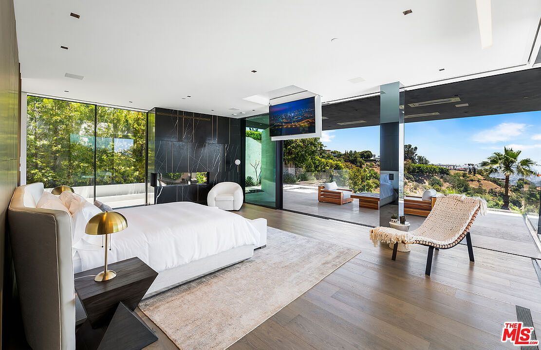This is a luxurious primary bedroom featuring floor-to-ceiling windows with views of lush greenery and the city. The room includes a king-size bed with white linens, a modern fireplace, and a retractable television. The hardwood floors and minimalist decor contribute to the room's contemporary and serene atmosphere.