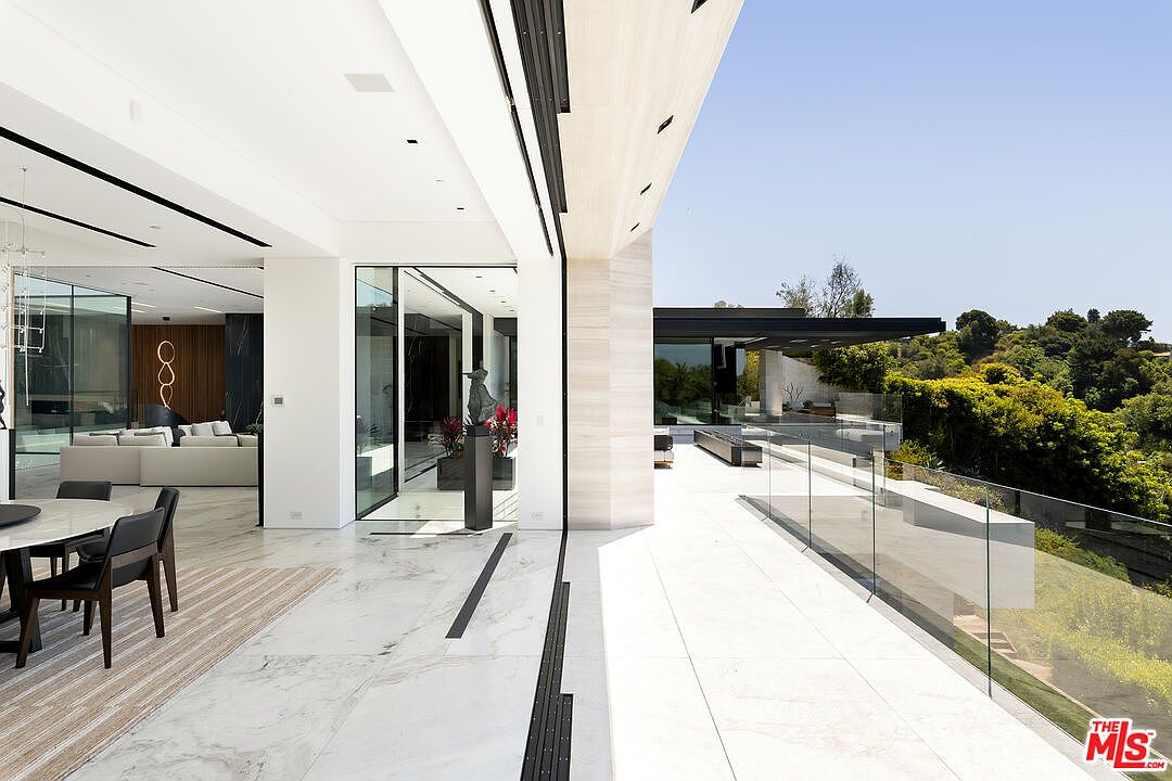 This image showcases a luxurious patio/balcony area seamlessly connected to the interior of a modern home. The space features sleek glass railings, light-colored tile flooring, and an open layout that extends to an outdoor lounge area with views of lush greenery. The design emphasizes indoor-outdoor living with a focus on clean lines and high-end finishes.
