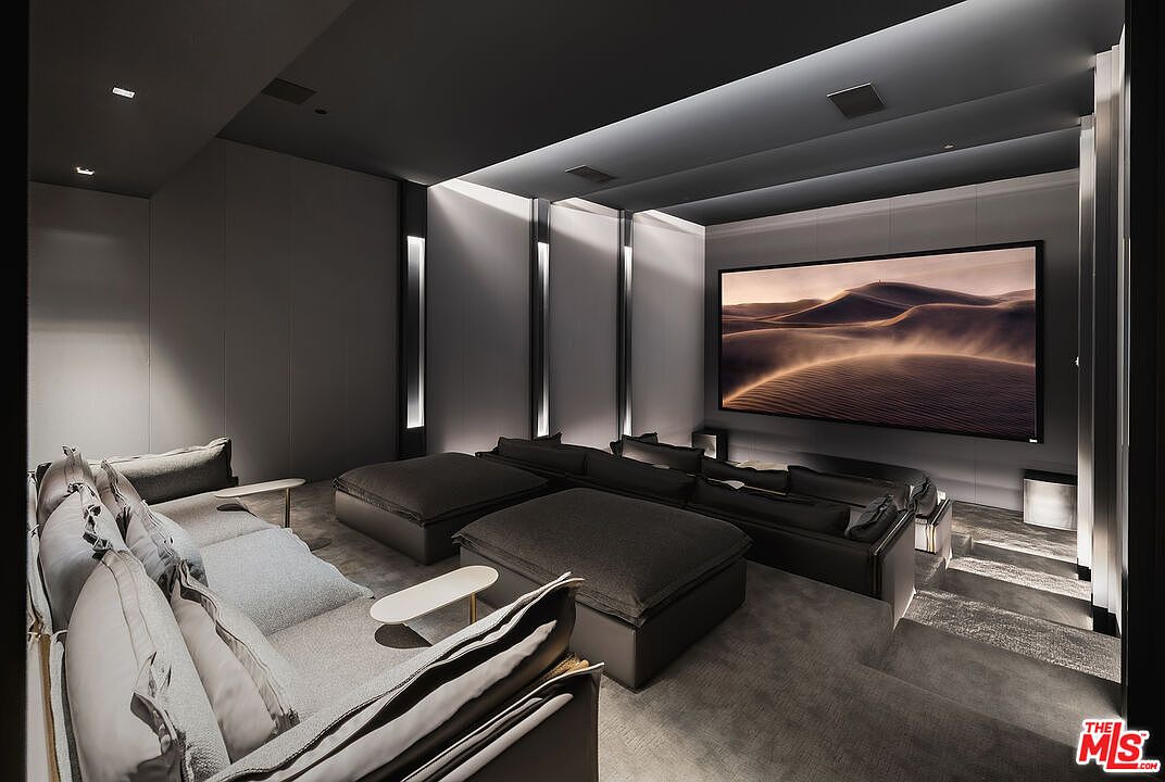 This is a modern home theater room featuring a large screen displaying a desert landscape. The room is furnished with multiple seating areas, including a long sofa and several ottoman-style seats, all in a dark, neutral color palette. The room has a sophisticated and luxurious feel, enhanced by the recessed lighting and minimalist design.