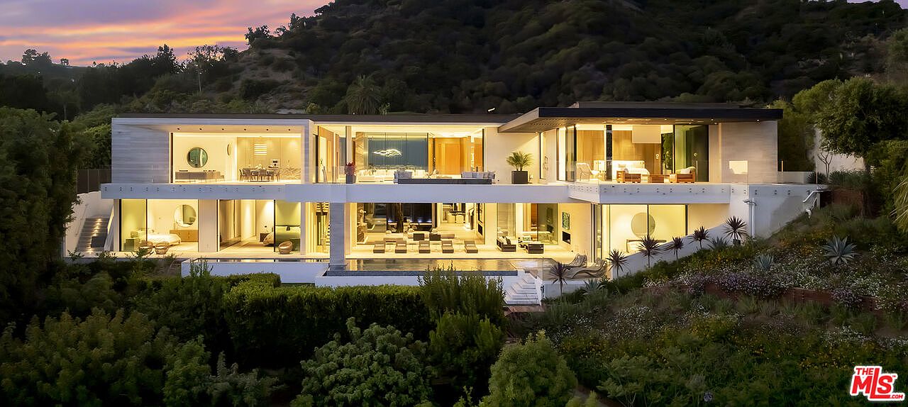 This is a rear view of a modern, multi-level luxury home. The house features expansive glass walls, allowing for seamless indoor-outdoor living. A pool is visible in the foreground, surrounded by lush landscaping, and the home is nestled into a hillside, providing privacy and stunning views.