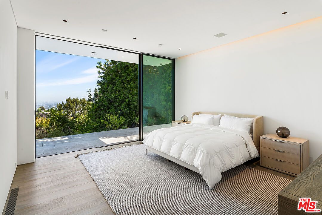This is a bright and airy primary bedroom featuring a large sliding glass door that opens to an outdoor patio with a view of lush greenery and the city skyline. The room is minimally decorated with a neutral color palette, a comfortable bed with white linens, and light wood nightstands. The overall impression is one of modern luxury and tranquility.
