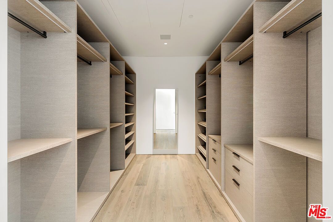 This is a well-organized walk-in closet featuring custom shelving and drawers on both sides of a central walkway. The closet is finished in a light wood tone, creating a clean and modern aesthetic. A doorway at the end of the closet provides access to another room, enhancing the sense of space and functionality.