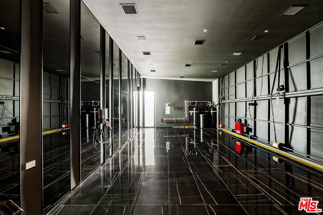 This is a modern garage interior featuring a sleek, dark aesthetic with black marble flooring and reflective wall panels. The space includes utility systems along the back wall and a door leading to another area. The overall impression is clean, minimalist, and high-end.