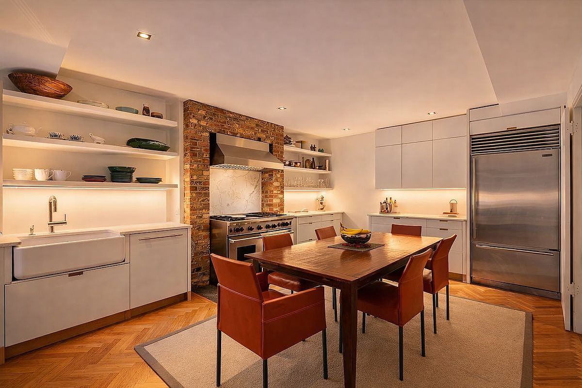 This sophisticated kitchen features a seamless blend of modern white cabinetry and rustic exposed brick, creating a warm and inviting atmosphere. A central wooden dining table serves as a focal point, complemented by vibrant orange chairs and a large stainless steel refrigerator. The space is illuminated by under-cabinet lighting and recessed ceiling lights, highlighting the herringbone hardwood floors and open shelving.