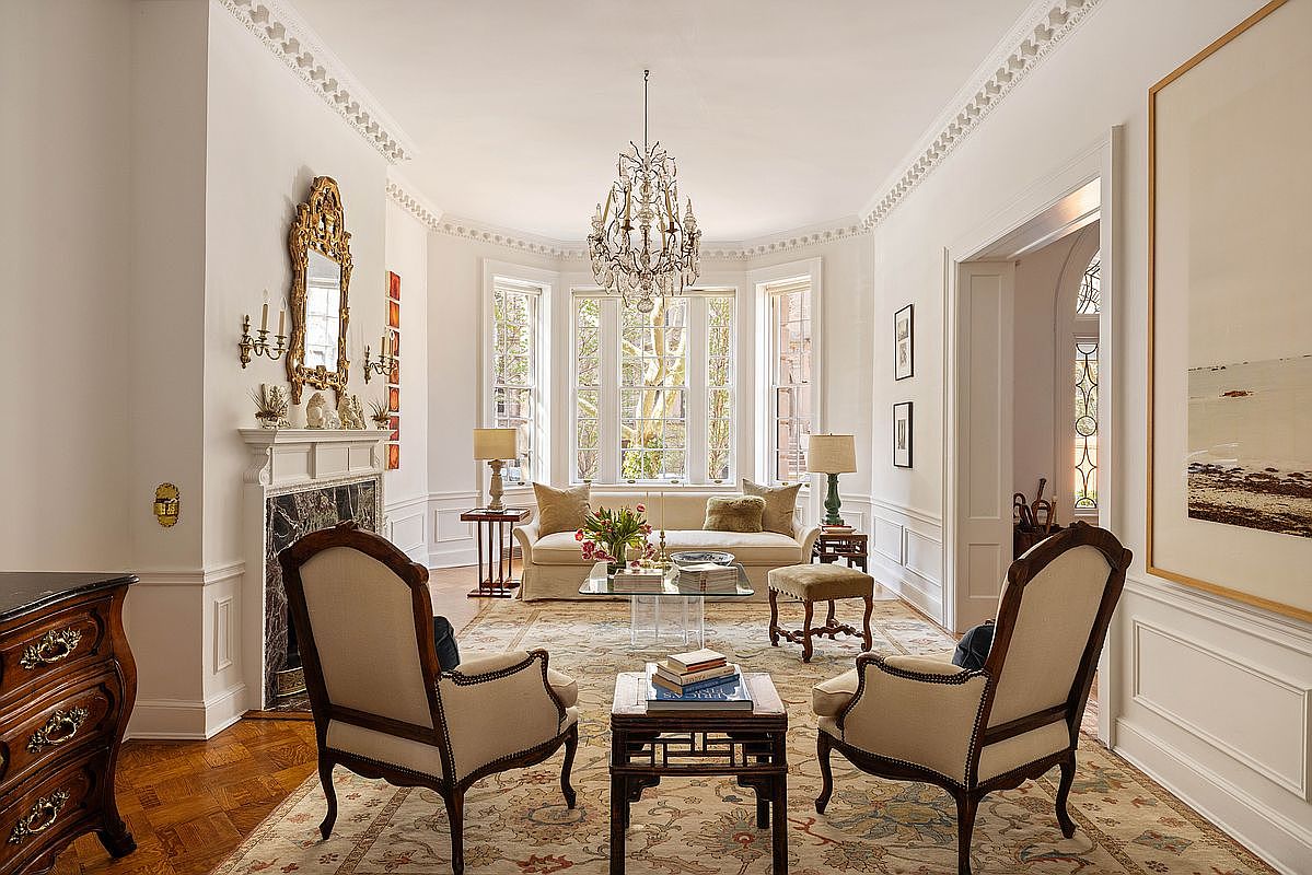 This elegant, formal living room features a sophisticated traditional design with a crystal chandelier, ornate crown molding, and a marble-surround fireplace. The space is anchored by a plush sofa and two classic armchairs arranged on a patterned area rug, creating a refined and inviting atmosphere. Large bay windows flood the room with natural light, highlighting the rich hardwood floors and the overall sense of luxury.