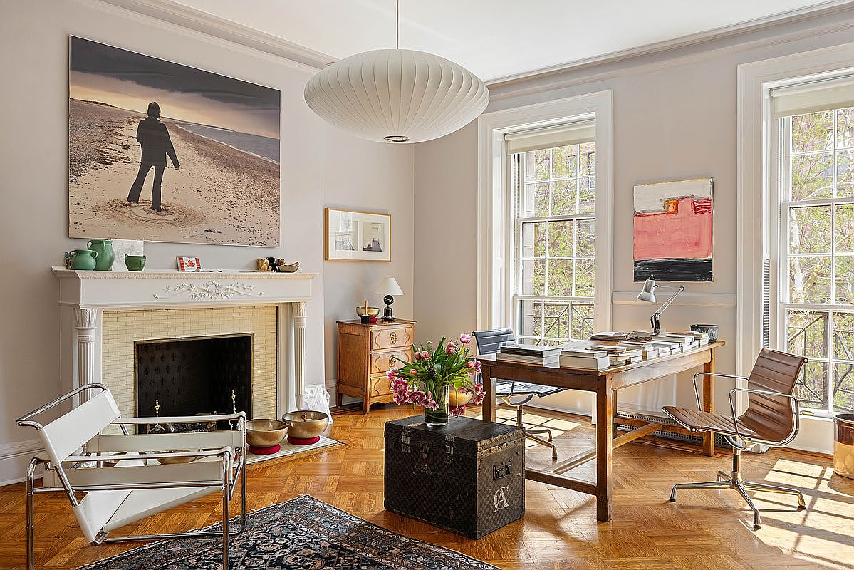 This sophisticated home office blends classic architectural details, such as a white fireplace mantel and herringbone hardwood floors, with modern design elements like a sleek desk and iconic mid-century modern chairs. A large, artistic photograph hangs above the fireplace, while a sculptural pendant light anchors the room, creating a bright and professional atmosphere. The space is bathed in natural light from two large windows, offering a serene and productive environment.