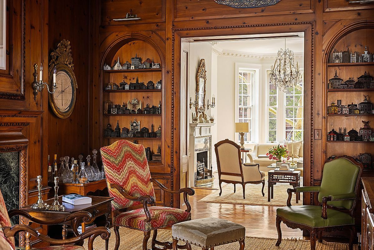 This elegant, wood-paneled study or living area exudes a classic, sophisticated atmosphere with its rich, warm-toned wood walls and built-in shelving filled with miniature architectural models. The room features a vibrant, patterned armchair, a green leather chair, and a small ottoman, all arranged around a side table, while an open doorway leads into a bright, sunlit sitting room with a fireplace and chandelier. The perspective is from within the study, looking through the threshold into the adjacent room, creating a sense of depth and refined luxury.