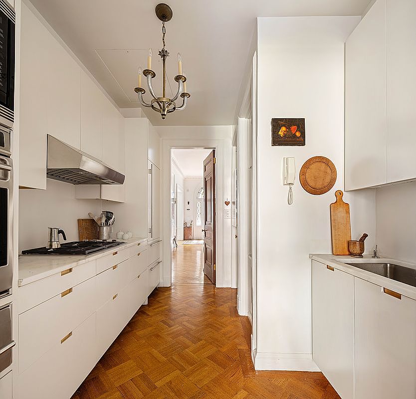 This galley-style kitchen features sleek, minimalist white cabinetry paired with warm, herringbone-patterned hardwood flooring. The space is illuminated by a classic brass chandelier, creating a sophisticated contrast between modern utility and traditional charm. A view through the doorway reveals a bright, inviting hallway, emphasizing the home's open and airy layout.